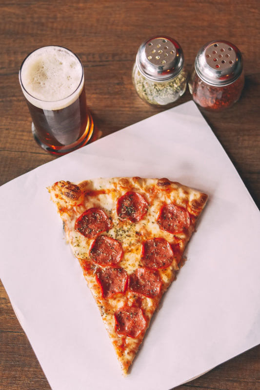 A beer, herb shakers and a slice of pizza sit on a table. Photo by Peter Bravo de los Rios via Unsplash